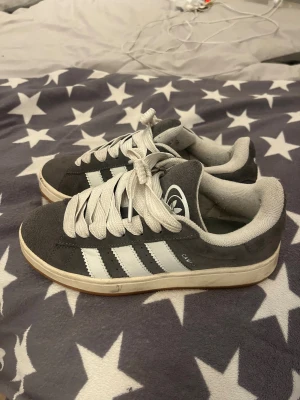 Adidas Campus grå sneakers mocka - Säljer ett par Adidas Campus sneakers i grå mocka med vita detaljer och klassiska tre ränder på sidan. Skorna har vit platt sula och breda vita snören. Perfekt för dig som gillar retrostil och vill ha ett par snygga och bekväma sneakers.