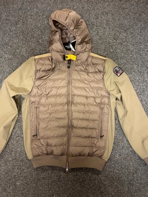 Beige pufferjacka från Parajumpers - Snygg beige pufferjacka från Parajumpers med huva och gul detalj vid halsen. Jackan har quiltad framsida, två dragkedjefickor och ribbade muddar. Patch på vänster ärm och stilren design som funkar till streetwear. Materialet är polyester och syntet.