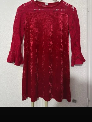 Röd sammetsklänning med spetsärmar - Supersnygg röd kortklänning i mjuk sammet med transparenta spetsärmar och volang vid ärmslut. Klänningen har rund halsringning och ett enkelt, rakt fall. Perfekt för dig som vill sticka ut med en elegant och trendig look.
