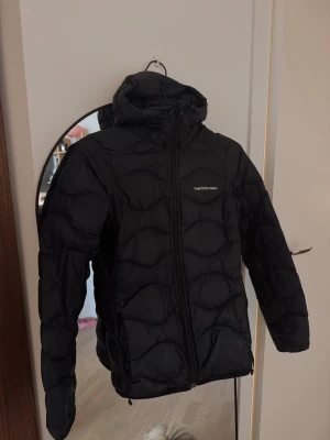 Peak Performance Helium Hood  - Svart dunjacka från Peak Performance i storlek XS. Jackan har huva, dragkedja framtill och quiltad design med vågiga sömmar. Perfekt för kalla dagar och har en clean look som funkar till det mesta. Kan va i behov av en tvätt då den legat nerpackad och är lite skrynklig. Annars i fint skick då den knappt blivit använd! 