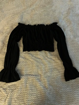 Svart offshoulder croppad blus - Säljer en svart blus med offshoulder och långa, voluminösa ärmar. Toppen är croppad med smockad midja och har en snygg rynkad struktur. Perfekt för dig som gillar trendiga och feminina detaljer.