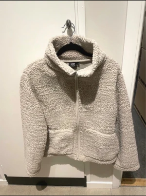 Beige teddyjacka från 157, storlek S - Mysig beige teddyjacka från 157 med dragkedja framtill och två stora fickor. Jackan har hög krage och är tillverkad i fluffigt teddy-material som håller dig varm. Perfekt för lager-på-lager och chill dagar.