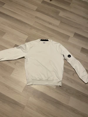 Vit sweatshirt från C.P. Company - Vit sweatshirt från C.P. Company med rund hals och lång ärm. Klassisk design med logotyp-patch på vänster ärm. Mjuk bomullskvalitet och ribbade muddar vid ärmslut och nederkant. Perfekt för en clean och stilren look. Kan ta emot byten👍Ekta✅