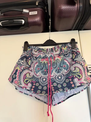 Mönstrade shorts från Odd Molly - Färgglada shorts från Odd Molly med paisley- och blommönster i blått, rosa, gult och vitt. Modellen har elastisk midja med rosa snörning och är tillverkad i 100% polyester. Perfekta för varma sommardagar när du vill sticka ut lite extra. Från märket Odd Molly. Storlek 3 som enligt deras storleksguide motsvarar en L. Men dessa kanske kan vara små i storleken. Mycket fint skick, jag hittar inga synliga anmärkningar eller defekter. 