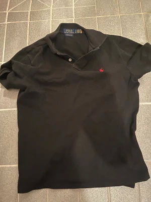 Svart Polo Ralph Lauren t-shirt slim fit - Svart t-shirt från Polo Ralph Lauren med klassisk krage och två knappar vid halsen. Custom slim fit-modell med röd broderad logga på bröstet. Tillverkad i mjuk bomull, perfekt för dig som gillar stilren och enkel design.