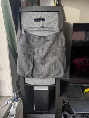 Grå kortkjol från Mexx med fickor - Snygg grå kortkjol från Mexx med bälteshällor och stora fickor med knappar framtill. Kjolen har en klassisk rak passform och är tillverkad i ett mjukt material som känns lite som ull. Perfekt för dig som gillar stilrena och praktiska plagg.