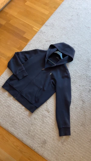 Mörkblå hoodie från Polo Ralph Lauren - Snygg mörkblå hoodie från Polo Ralph Lauren med dragkedja, huva och kängurufickor. Klassisk logga broderad på bröstet. Tillverkad i mjuk bomullsblandning som är skön att ha på sig. Perfekt för en chill och stilren look.