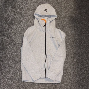 Grå hoodie från Sail Racing med huva - Snygg ljusgrå hoodie från Sail Racing med svart dragkedja och logga på bröstet samt huvan. Tröjan har långa ärmar, fickor framtill och är tillverkad i mjuk bomullsmix som är perfekt för kyliga dagar. Enkel och stilren design som passar till jeans eller mjukis. 150 storlek