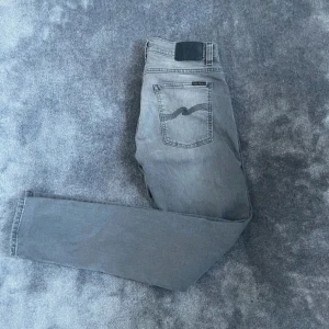 Nudie Jeans Gråa Slim Fit  - Snygga och bekväma Nudie Jeans i modellen Tight Terry (Slim Fit) i storlek W32 L32. Färg: Grå, vilket gör dem enkla att matcha både till vardags och lite mer uppklädda tillfällen. Jeansen är i mycket fint skick och har endast använts varsamt.  🔹 Märke: Nudie Jeans 🔹 Modell: Tight Terry – Slim Fit 🔹 Storlek: W32 L32 🔹 Färg: Grå 🔹 Skick: Mycket fint skick  ⸻  Liknande märken: Levi’s, Acne Studios, Neuw, Dr. Denim, Cheap Monday  Sökord: nudie jeans herr, tight terry jeans, slim fit jeans grå, nudie