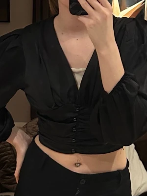 Svart croppad blus med knappar - Säljer en svart croppad blus med djup v-ringning och puffiga långärmar. Toppen har snygga knappar framtill och är gjord i ett glansigt, mjukt material. Perfekt för lite finare tillfällen! Från Missay storlek XS