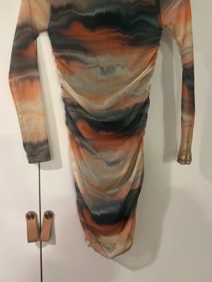 Randig meshklänning från Gina Tricot - Snygg meshklänning från Gina Tricot med långa ärmar och coolt randigt mönster i orange, beige, svart och grå toner. Klänningen är kort och har en figurnära passform med lätt rynkade detaljer längs sidorna. Perfekt för dig som gillar trendiga och färgstarka plagg.