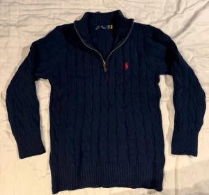 Blå stickad tröja från Polo Ralph Lauren - Kabelstickad blå tröja från Polo Ralph Lauren med halv dragkedja i halsen och klassisk röd logga broderad på bröstet. Tröjan har lång ärm och ribbade muddar för en snygg passform. Perfekt för lager-på-lager under kyliga dagar.