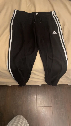 Svarta Adidas mjukisbyxor med vita ränder - Svarta mjukisbyxor från Adidas med klassiska vita ränder längs sidorna och logga på vänster ben. Byxorna har snörning i midjan och är tillverkade i mjukt material som är perfekt för chill eller träning. Passformen är loose och muddar vid bensluten.