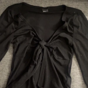 Svart knytblus från Gina Tricot XS - Svart långärmad blus från Gina Tricot i storlek XS. Toppen har en djup v-ringning och snygg knytning framtill som ger en trendig look. Tillverkad i mjukt material som sitter skönt på kroppen. Perfekt att styla med jeans eller kjol.