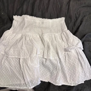 Vit volangkjol från Kappahl - Supersöt vit kortkjol från Kappahl med volanger och diskret prickigt mönster. Kjolen har smock i midjan för en bekväm passform och är gjord i ett lätt bomullstyg. Perfekt för en fräsch och somrig look!