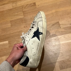 Golden Goose Superstar sneakers vit/svart - Golden Goose Superstar sneakers i vitt skinn med svart stjärndetalj på sidan och svart hälkappa. Klassisk låg modell med snörning och rund tå. Snygg vintage-look och ikonisk branding på hälen. Perfekta för dig som gillar streetstyle och exklusiva detaljer.