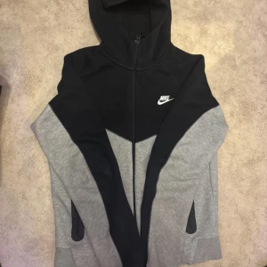 Nike hoodie svart och grå S - Nike hoodie i svart och grått med vit logga på bröstet. Tröjan har hel dragkedja, huva och två fickor med dragkedja. Materialet är 53% bomull och 47% polyester, vilket gör den mjuk och skön. Perfekt för chill eller träning. Som ny inte ny dock.
