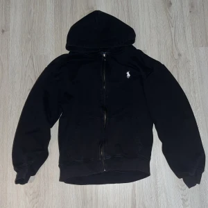 Svart hoodie från Polo Ralph Lauren - Svart hoodie från Polo Ralph Lauren med vit broderad logga på bröstet. Tröjan har huva, dragkedja framtill och två fickor. Tillverkad i mjuk bomull och har ribbade muddar vid ärmslut och nederkant. Perfekt för chill dagar och streetstyle.