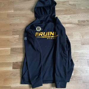 Svart Bruins hoodie från Fanatics - Svart hoodie från Fanatics med Boston Bruins-logga och NHL-märke på ärmen. Tröjan har huva, långa ärmar och tryck i gult och vitt framtill. Perfekt för hockeyfans som vill visa sitt lagstöd. Materialet är mjukt och känns sportigt.