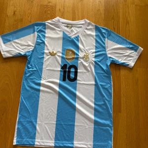 Argentina Messi #10 fotbollströja Adidas storlek M - Säljer en klassisk Argentina fotbollströja med Messi och nummer 10 på ryggen. Tröjan har breda blå och vita ränder, guldiga detaljer och FIFA-märke på bröstet. Tillverkad i lätt och ventilerande material, perfekt för match eller träning.