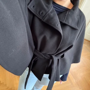 Svart capejacka med bälte XS/S - Svart capejacka från Reserved med bred krage och knytbälte i midjan. Jackan har en loose passform och är tillverkad i mjukt material som ger en snygg siluett. Perfekt för dig som gillar stilrena och trendiga plagg med lite edge.