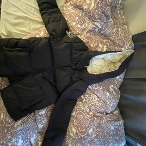 Svart pufferjacka från db, stl S - Svart pufferjacka från db med broderad logga på ärmen. Jackan har huva, stora fickor och dragkedja framtill. Insidan är ljus och jackan är vadderad för extra värme. Perfekt för kalla dagar när du vill hålla stilen.