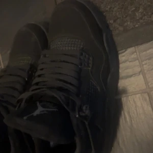 Svarta Nike Jordan 4 sneakers - Säljer ett par helsvarta Nike Jordan 4 sneakers med klassisk Jumpman-logga på plösen. Skorna har snörning, mesh-detaljer och en chunky sula som ger en sportig vibe. Perfekta för dig som gillar streetstyle och vill ha en ikonisk look.