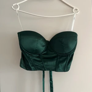 Mörkgrön satin korsett från NA-KD - Supersnygg mörkgrön korsett från NA-KD i glansigt satinmaterial. Toppen har bygelkupor, smala axelband och snörning i ryggen för perfekt passform. Perfekt att styla med jeans eller kjol för en trendig look.