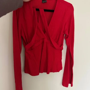 Röd omlottopp med v-ringning - Snygg röd omlottopp med djup v-ringning och långa ärmar. Toppen har en cool omlottdetalj framtill och är gjord i ett lätt, strukturerat tyg som ger en trendig look. Perfekt att styla med jeans eller kjol för en uppdaterad outfit.