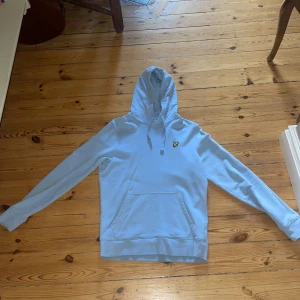 Ljusblå hoodie från Lyle & Scott - Ljusblå hoodie från Lyle & Scott med klassisk gul broderad logga på bröstet. Tröjan har huva med snörning, magficka och långa ärmar. Perfekt för dig som gillar en clean och stilren look.