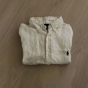 Vit linneskjorta från Ralph Lauren - Vit skjorta från Ralph Lauren i luftig linne med klassisk krage och knappar framtill. Ikonisk svart broderad logga på bröstet. Perfekt för en clean och stilren look.