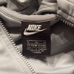 Ljusgrå Nike hoodie med dragkedja XS - Nike hoodie i ljusgrått med hel dragkedja och huva. Snygg logga på bröstet och Nike-band längs ärmarna. Tillverkad i mjuk polyester, perfekt för chill eller träning. Stilsäker och sportig vibe.