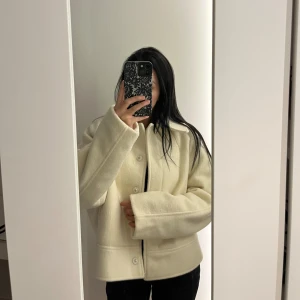 Vit ulljacka med knappar - Snygg vit/ beige liknande jacka från Gina Tricot i ullblandning. Jackan har en rak, boxig passform med stora knappar framtill, klassisk krage och diskreta fickor på sidorna. Perfekt för lager-på-lager och ger en clean look till alla outfits. 