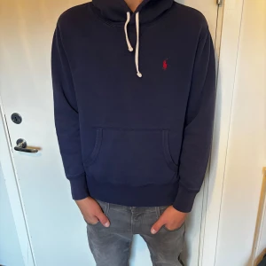Mörkblå hoodie från Polo Ralph Lauren - Mörkblå hoodie från Polo Ralph Lauren med vit snörning och klassisk röd logga broderad på bröstet. Tröjan har känguruficka framtill, huva och långa ärmar. Tillverkad i mjuk bomull för en skön och avslappnad stil. Priset kan såklart diskuteras