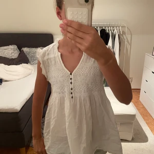 Vit Odd Molly tröja med spets - Supersöt vit klänning från Odd Molly med v-ringning, knappar framtill och fina spetsdetaljer upptill. Ärmlös modell med volang vid axlarna och luftig passform. Perfekt för varma dagar och ger en romantisk vibe.