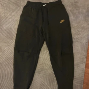 Svarta mjukisbyxor från Nike, M - Svarta mjukisbyxor från Nike med guldfärgad logga på vänster ben. Byxorna har elastisk midja med snörning och dragkedjeförsedda fickor. Sköna och stilrena, perfekta för chill eller träning. Tillverkade i mjukt bomullsmaterial. Knappt använda.