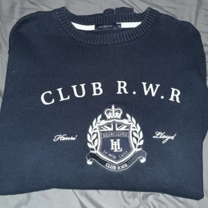Henri Lloyd tröja  - Mörkblå stickad tröja från Henri Lloyd med broderad Club R.W.R-logga och emblem på bröstet. Klassisk rund halsringning och ribbade muddar. Perfekt för dig som gillar preppy stil och snygga detaljer.och även lite lyxigare stil.