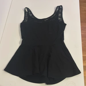 Svart peplum-linnetopp från Gina Tricot - Säljer en svart peplum-linnetopp från Gina Tricot i storlek M. Toppen har breda axelband och v-ringning med snygga spetsdetaljer upptill. Modellen är ärmlös och har en utställd nederdel som ger en fin siluett. Perfekt att styla med jeans eller kjol.
