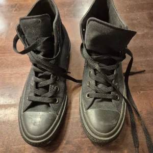 Svarta Converse All Star high-top - Klassiska svarta Converse All Star high-top sneakers i canvas med svarta snören och gummisula. Skorna har rund tå, platt sula och metallöljetter. Har lunarlon sula som är mjukare än den vanliga sulan. Perfekta för en cool och avslappnad stil. Unisexmodell, storlek 39 (24,5 cm). Använda någon enstaka gång.