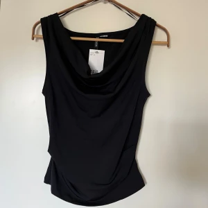 Svart draperad top från HM - Säljer en svart ärmlös topp från H&M i storlek S. Toppen har  jättesnygg draperad halsringning och är figurnära i modellen. Oanvänd. Passar s/m m, stretchig i tyget.