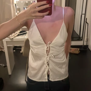 Vit satintopp med snörning - Säljer en vit satintopp med djup v-ringning och smala axelband. Toppen har snygg snörning framtill som ger en trendig look. Perfekt för dig som gillar stilrena och feminina plagg med det lilla extra.