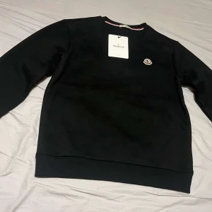 Svart sweatshirt från Moncler - Svart sweatshirt från Moncler med rund hals och klassisk logotyp på bröstet. Tröjan har ribbade muddar och en snygg detalj med röd, vit och blå rand på insidan av kragen. Perfekt för dig som gillar stilrena märkesplagg.