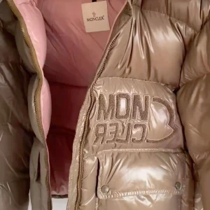 Moncler brun pufferjacka med rosa detaljer - Snygg pufferjacka från Moncler i glansig brun nyans med rosa foder och axeldetaljer. Jackan har broderad Moncler-logga på bröstet, stora fickor med knappar och dragkedja framtill. Perfekt för dig som vill sticka ut med en lyxig och trendig vinterjacka. Helt nytt kommer med förpackning 