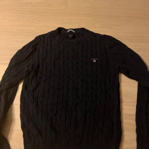 Svart kabelstickad tröja från GANT - Kabelstickad svart tröja från GANT i storlek S. Klassisk rund halsringning, ribbade muddar och broderad GANT-logga på bröstet. Perfekt för dig som gillar stilrena och tidlösa plagg med snygg struktur. Priset kan sänkas