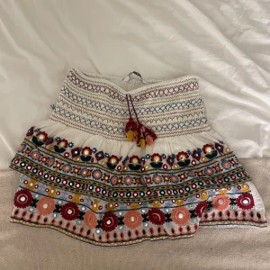 Broderad kortkjol från Zara XS - Superfin kortkjol från Zara i vitt med färgglada broderier och spegeldekorationer. Kjolen har smockad midja, snörning med tofsar och blommigt mönster i rött, blått, gult och grönt. Perfekt för dig som gillar boho-vibes och detaljer!