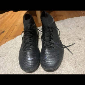 Svarta Nike Mercurial fotbollsskor - Säljer ett par svarta Nike Mercurial fotbollsskor med snörning och högre skaft. Skorna har en snygg, diskret glansig finish och klassisk Nike Swoosh på sidan. Utrustade med dobbar för gräsplan och tillverkade i syntetmaterial för bästa grepp och känsla på planen.