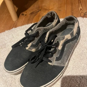 Vans sneakers med camomönster - Säljer ett par Vans sneakers med svart mocka framtill och klassiskt camouflagemönster på sidorna. Skorna har svarta snören, vit sula och den ikoniska vågformen på sidan. Perfekta för dig som gillar streetstyle och vill sticka ut lite extra.