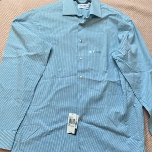 Blå randig skjorta från Calvin Klein - Snygg blå skjorta från Calvin Klein med tunna vita ränder och klassisk krage. Regular fit och långärmad modell med knappar framtill. Perfekt för dig som gillar stilrena och tidlösa plagg. Tillverkad i mjuk bomull för en skön känsla hela dagen.