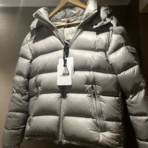 Grå pufferjacka från Moncler - Säljer en grå pufferjacka från Moncler med huva och dragkedja framtill. Jackan har fickor med dragkedja och Moncler-logga på ärmen. Perfekt för kalla dagar och riktigt snygg till streetwear-stilen. Jackan är i storlek 3. Jackan har såklart även en fungerande NFC tag som tar en till monclers hemsida 