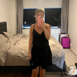 Svart kort klänning med tunna axelband - Säljer en svart kort klänning med tunna axelband och v-ringning. Klänningen har en luftig passform och faller snyggt med lätt volang nedtill. Perfekt för varma dagar och enkel att styla med smycken eller en jeansjacka.